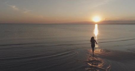 Aerial forward fly over teen girl barefoot running into water on baltic sea beach and raising arms in sunset time