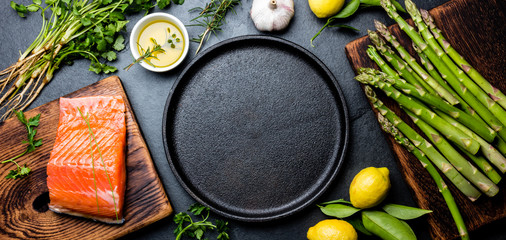 Raw salmon fillet, asparagus, lemons and herbs around cast iron plate. Food cooking background with...
