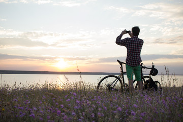 silhouette of a man with touring road bike watching and make photo of sunset in lake on cellphone 