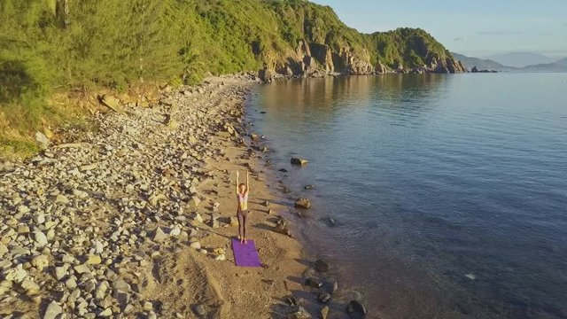 Drone Rises over Girl Standing in Yoga Pose on Beach by Hill