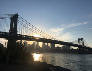 Sunset sets over the Manhattan Bridge and the East River, view from Brooklyn shore. Manhattan Bridge in silhouette against setting sun, New York Financial district and skyscrapers at sunset.
