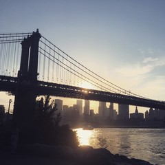 View of the Manhattan Bridge at sunset from Brooklyn shoreline. Looking towards New York Financial District at sunset, Manhattan Bridge spans the East River as the sun sets.