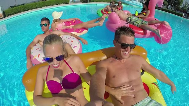 SLOW MOTION SELFIE: Smiling young couple with friends laughing on colorful floaties in pool. Happy teenagers enjoying summer vacation on inflatable pizza, flamingo, watermelon and doughnut floats