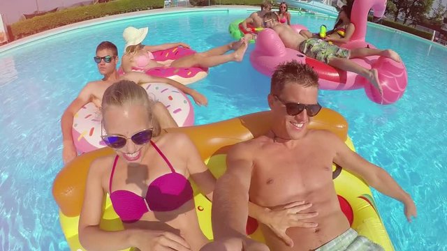 SLOW MOTION SELFIE LENS FLARE Smiling young couple with friends on colorful floaties in pool. Happy teenagers laughing and enjoying summer vacation on inflatable pizza, flamingo and doughnut floats