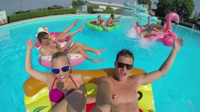 SLOW MOTION SELFIE: Smiling young couple with friends enjoying on colorful floaties in pool. Happy teenagers having summer water fight on inflatable pizza, flamingo, watermelon and doughnut floats