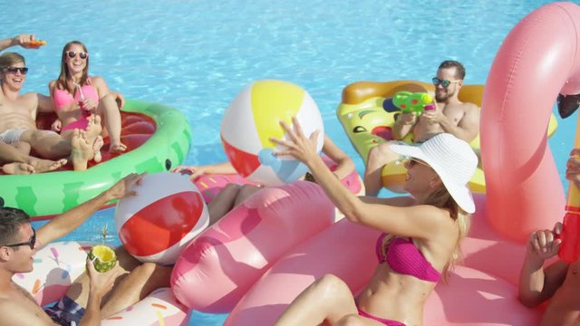 SLOW MOTION CLOSE UP Happy smiling students have water gun fight on colorful floaties at pool party on spring break. Cheerful young people have fun on inflatable flamingo pizza watermelon and doughnut