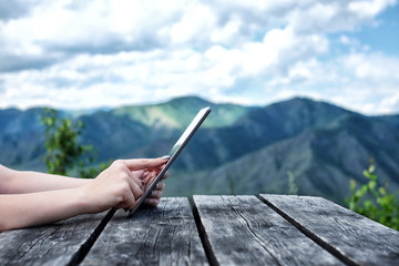 Woman's hands using tablet pc sitting at the old wooden table among the mountains.