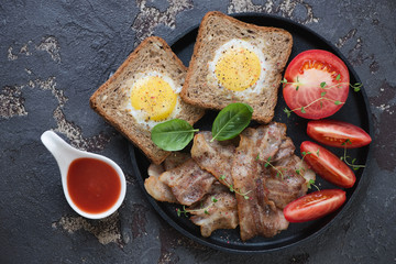 Eggs in the basket served with fried bacon and tomato on a metal plate, above view on a brown stone background