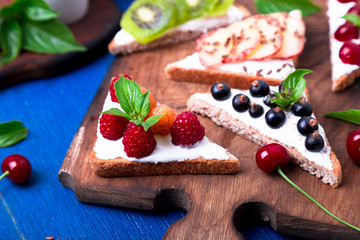 Fruit toast on wooden board on blue rustic background. Healthy breakfast. Clean eating. Dieting concept. Grain bread slices with cream cheese and various fruit, berries, seeds. Vegetarian.