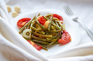 Baked vegetables with silver fork on a white plate cooked in the oven - bean, potato, onion, tomato. On a white surface. Decorated with garlic. Side view