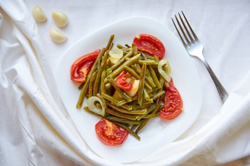 Baked vegetables - bean, potato, onion, tomato on a white plate cooked in the oven. On a white surface. Decorated with fresh garlic and silver fork. Top view