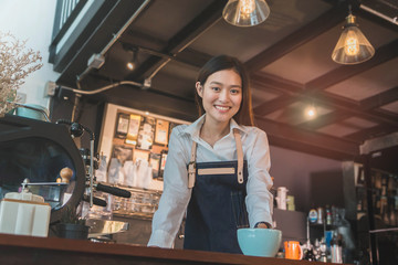 Young beautiful Asian woman barista wear blue apron holding hot coffee cup served to customer at bar counter in coffee shop with smile face.Concept of cafe and coffee shop small business.Vintage tone