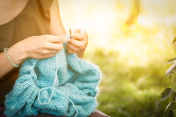 A young woman in a summer green top and skirt stands in a birch forest on a summer day and knits a blue sweater from natural mohair threads, in the background a green sun shines