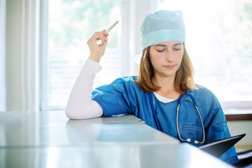 Young female thoughtful medical doctor in a blue uniform standing indoors in a clinic, leaning on a metal filing cabinet, holding a pen and looking into the folder.