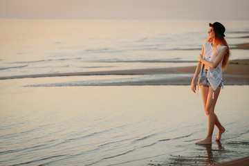 Young, beautiful girl walking at the beach at sunset. Stylish woman with long hair standing in blouse and jeans shorts