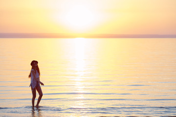 Young, beautiful girl walking at the beach at sunset. Stylish woman with long hair standing in blouse and jeans shorts