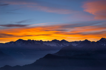 Colorful sunlight behind majestic mountain peaks of the Italian - French Alps, viewed from distant. Fog and mist covering the valleys below, autumnal landscape, cold feeling.