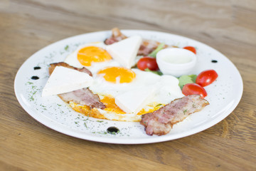 Fried eggs with bacon on the wooden table. Tomato and cheese on the side.