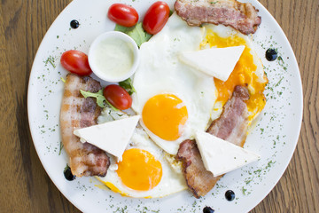 Fried eggs with bacon on the wooden table. Tomato and cheese on the side.