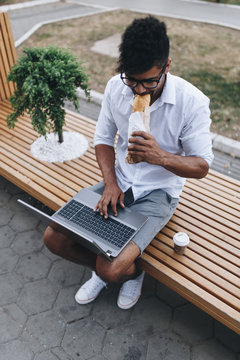 Young handsome Afro American man with glasses sitting on street with laptop and eating sandwich. Business concept.