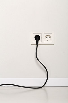 Black power cord cable plugged into european wall outlet on white plaster wall