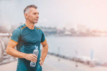 Portrait of healthy athletic middle aged man with fit body holding bottle of refreshing water,...