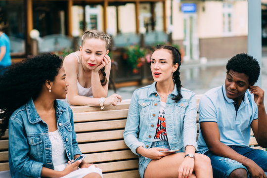 Group Of Four Multiethnic Hipster Tenager Friends Playing Guitar, Singing And Sitting On The Bench Outdoors In City Square.