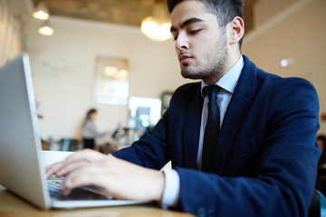 Serious young businessman typing on laptop keypad in cafe