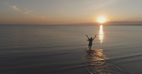 Aerial shot over teen girl barefoot running and raising arms in water on baltic sea beach in sunset time