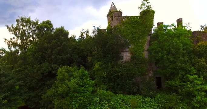 Aerial view of derelict castle ruins overgrown with trees with Loch Lomond and mountains in the distance.