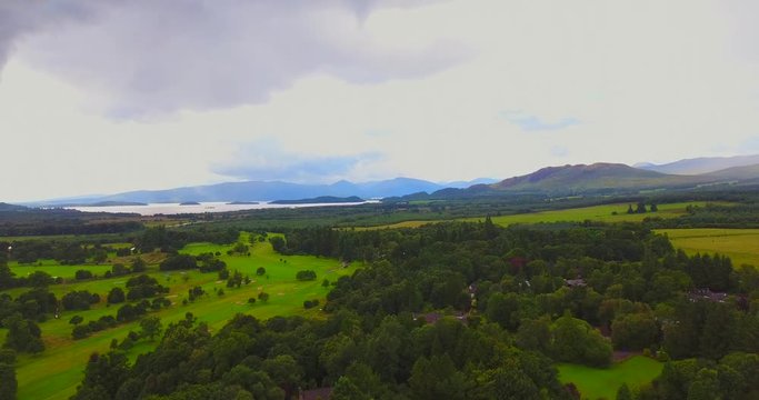Aerial view of derelict castle ruins overgrown with trees with Loch Lomond and mountains in the distance.