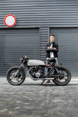 Handsome rider man with crossed arms in black biker jacket near classic style cafe racer motorcycle industrial gates as background. Bike custom made in vintage garage. Brutal fun urban lifestyle. - 166894425