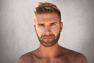 Close up portrait of good-looking male with dark eyes, bristle and trendy hairdo being naked, looking with serious expression at camera, isolated over grey background. Topless young macho man