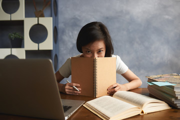 Naklejka premium Charming Chinese student girl studying in modern library, working on research project for course paper, sitting at desk with laptop, open book, covering her mouth with copybook and looking at camera