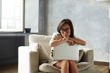 Obraz premium Pretty girl of Asian appearance in stylish eyeglasses relaxing on comfortable armchair in light spacious room with large window, holding generic laptop computer, looking away with dreamy expression