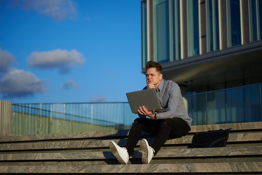Thoughtful young European creative worker sitting on concrete stairs outdoors, holding laptop computer in his hands, working on project, deep in thoughs. People, creativity and modern technologies