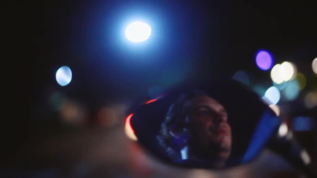 Young handsome extreme man drives the motorcycle in night reflected in mirror on blurred bokeh light night beautiful background. 1920x1080