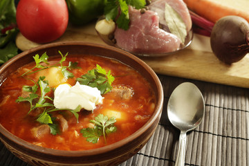 Clay brown plate and spoon with red borsch with sour cream on a table near vegetables and meat