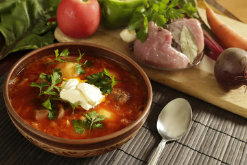 Clay brown plate and spoon with red borsch with sour cream on a table near vegetables and meat