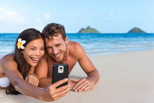 Beach vacation couple taking fun phone selfie on Hawaii vacation. Asian girl Caucasian man relaxing on Lanikai beach, Oahu, on summer holidays using smartphone for pictures together.