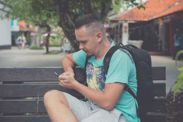 Happy young handsome man sitting on the bench outdoors and using smartphone. Tropical island of Bali, Indonesia.