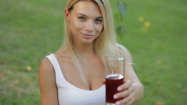Healthy lifestyle drink. Young woman give a garnet juice sitting outdoors at meadow in slow motion