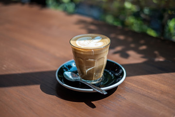 Cup of hot milk coffee with rosetta latte art with green tree leaves as background. Concept of morning drink freshness. Selective focus