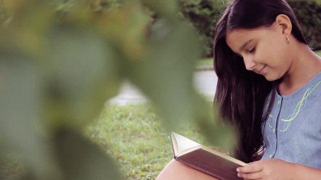 Portrait. A beautiful little girl with long hair reads a book sitting under a tree and dreams about something pleasant smiling