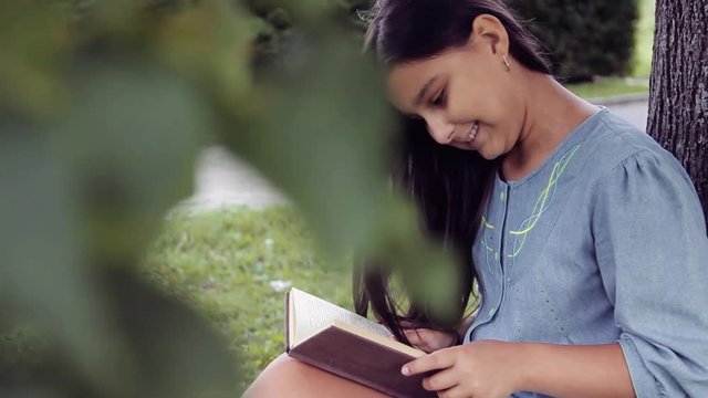 Portrait. A beautiful girl with long hair reads a book sitting under a tree and dreams about something pleasant smiling