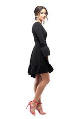 Fototapeta premium Side view of gorgeous female fashion model in black dress smiling at camera posing. Full body length portrait isolated on white studio background.