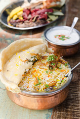 Authentic chicken biryani served with naan bread, fragrant pilau rice and yoghurt, in a metal pot.