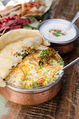 Authentic chicken biryani served with naan bread, fragrant pilau rice and yoghurt, in a metal pot.
