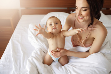 Funny nice little baby girl stretching hands to camera sitting on bed with mom at home.