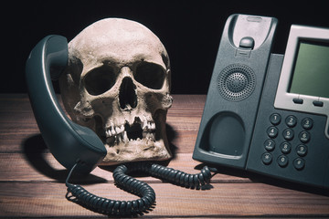 Office work concept. Human skull model with modern telephone on wooden table and black background.